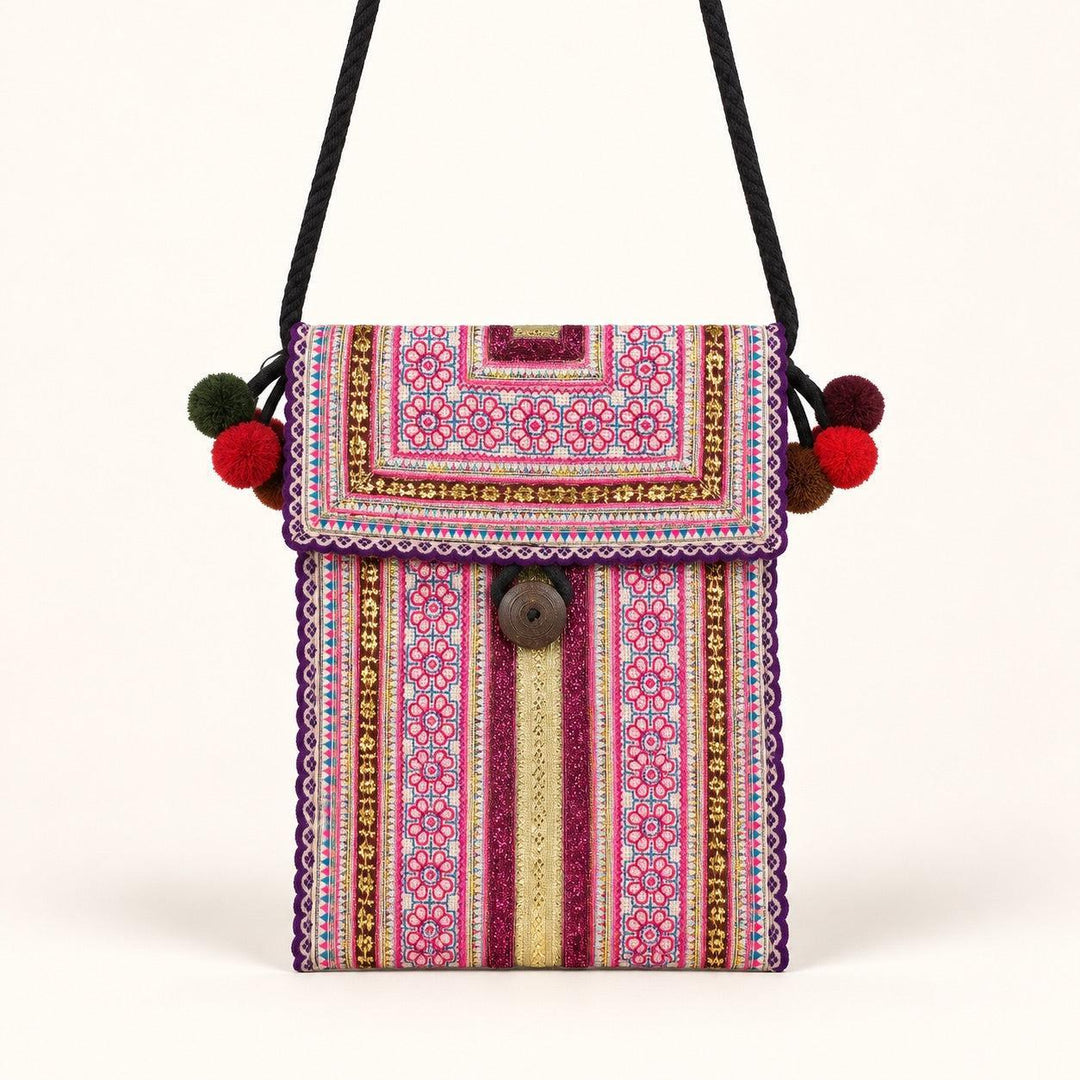 A small embroidered crossbody bag with a black strap, featuring a vibrant pink, red, and gold geometric pattern, a brown button closure, and colorful pom-poms on the strap ends, set against a plain off-white background.