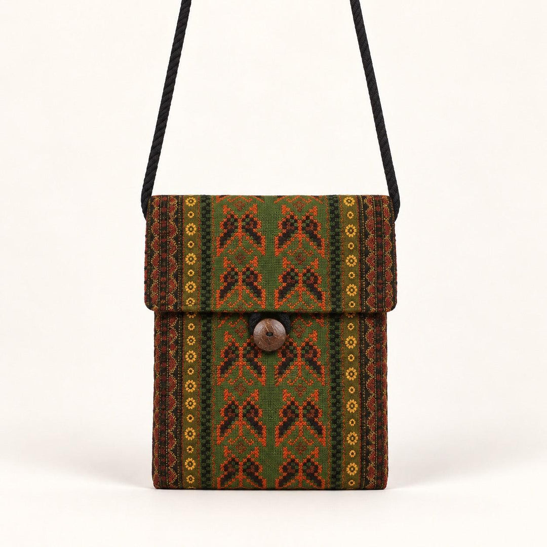 A small woven crossbody bag with a black strap, featuring a green, brown, and orange geometric pattern with a central wooden button closure, set against a plain off-white background.
