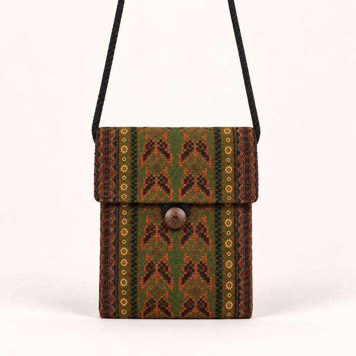 A small woven crossbody bag with a black strap, featuring a green, brown, and orange geometric pattern with a central wooden button closure, set against a plain off-white background.