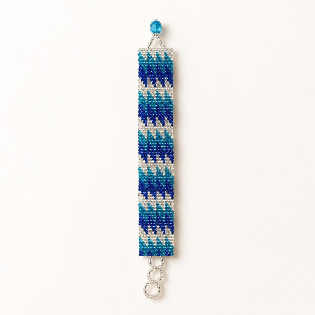 A handmade beaded bracelet featuring a geometric chevron pattern in shades of blue, teal, and white, with a silver-tone split ring at the bottom and a small blue bead at the top, shown against a plain white background.