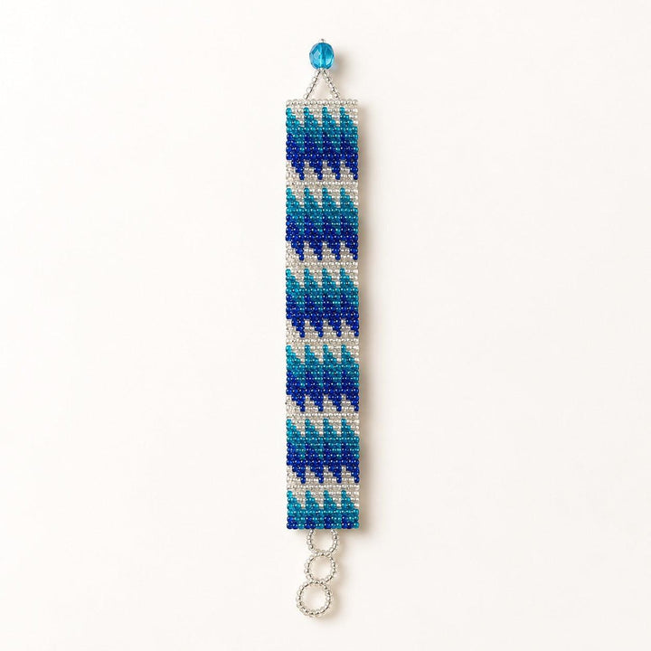 A handmade beaded bracelet featuring a geometric chevron pattern in shades of blue, teal, and white, with a silver-tone split ring at the bottom and a small blue bead at the top, shown against a plain white background.