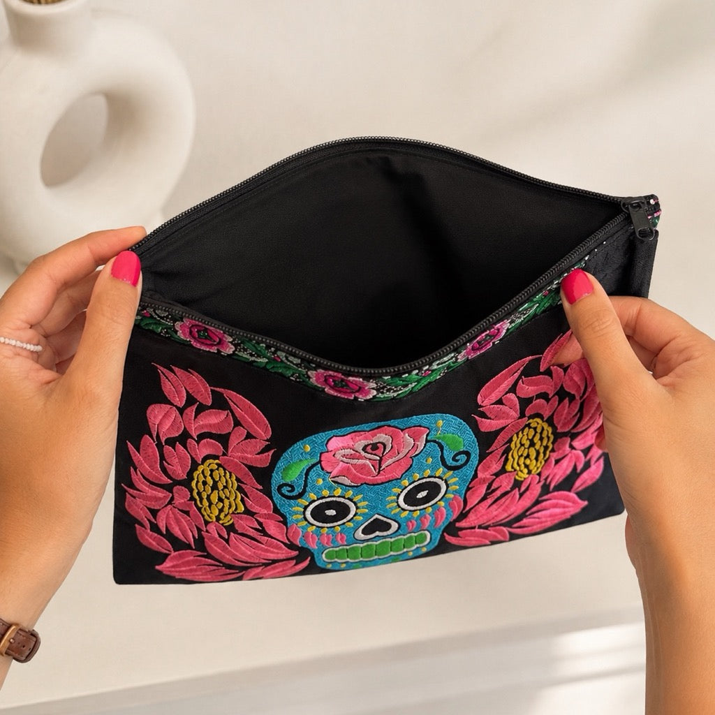 A black zippered cosmetic pouch with colorful floral and sugar skull embroidery, held open by two hands with pink nail polish against a light background.