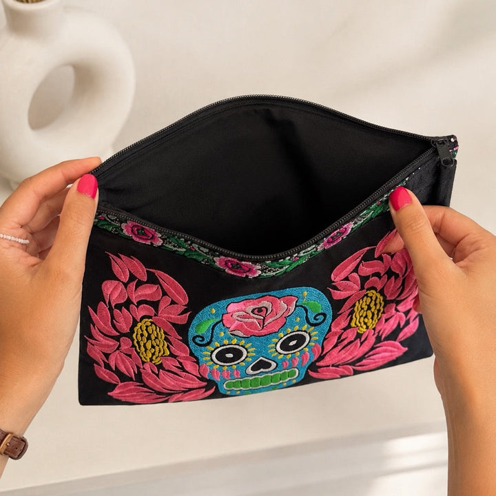 A black zippered cosmetic pouch with colorful floral and sugar skull embroidery, held open by two hands with pink nail polish against a light background.
