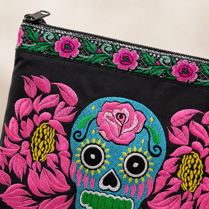 Black fabric clutch bag with colorful embroidered floral and sugar skull designs, featuring a pink rose and large pink flowers with yellow centers, and a green and pink floral border along the zipper.