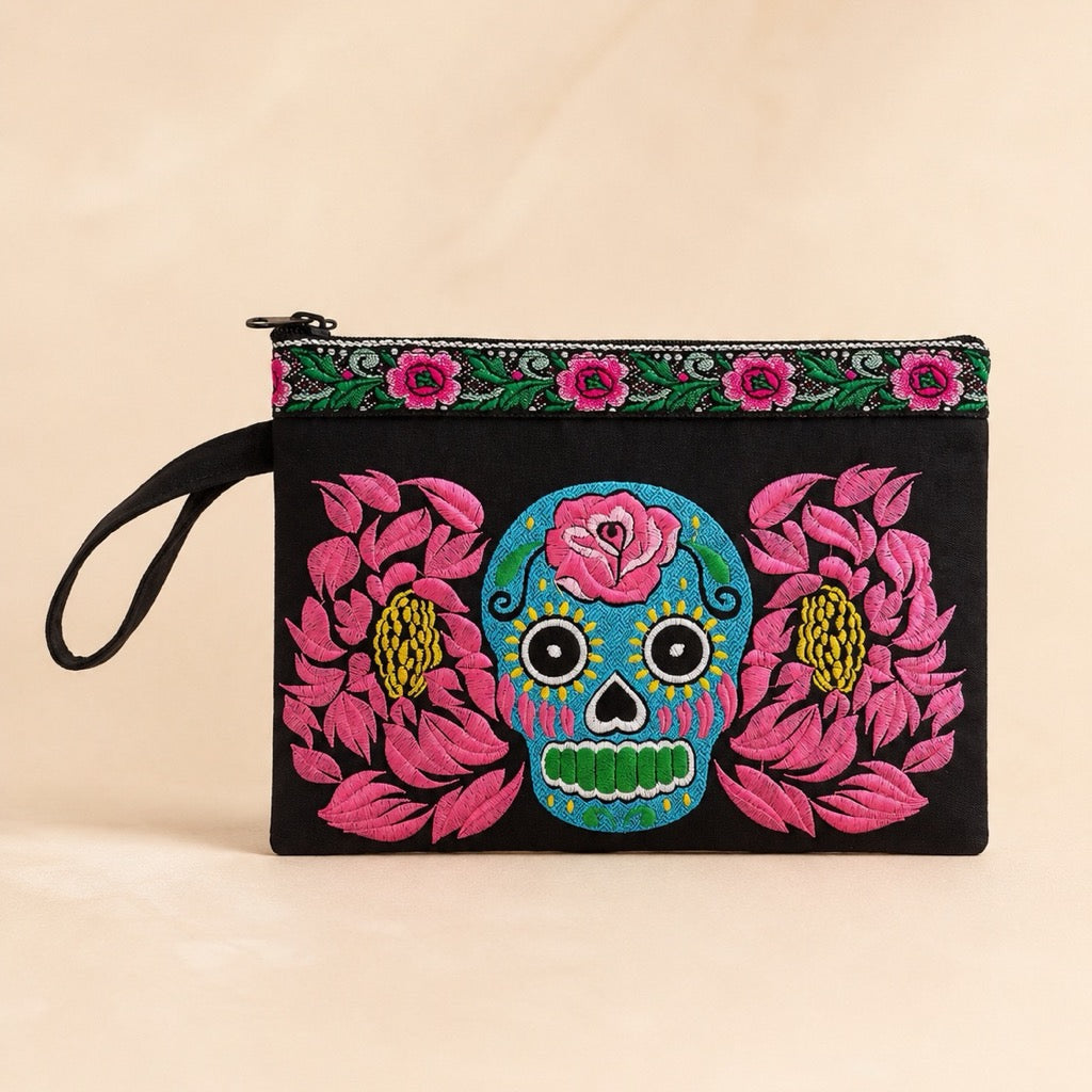 A black fabric clutch pouch with a Day of the Dead sugar skull and pink floral embroidery, featuring a top zipper and wrist strap, set against a warm beige background.