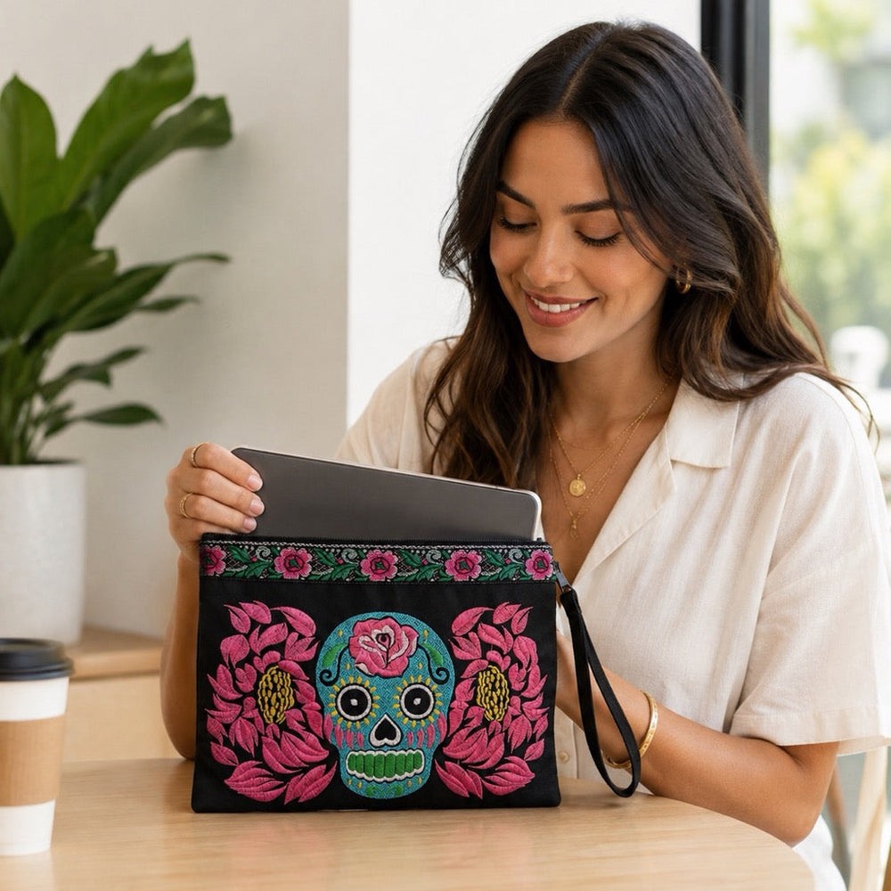 A woman in a white shirt sits at a wooden table holding a black sugar skull handmade clutch bag decorated with a colorful embroidered sugar skull and pink flowers. A coffee cup and a potted plant are visible in the background.