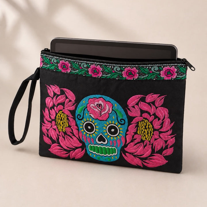Black clutch pouch with colorful embroidered floral and sugar skull designs, a wrist strap, and a zipper closure, shown on a light beige background.