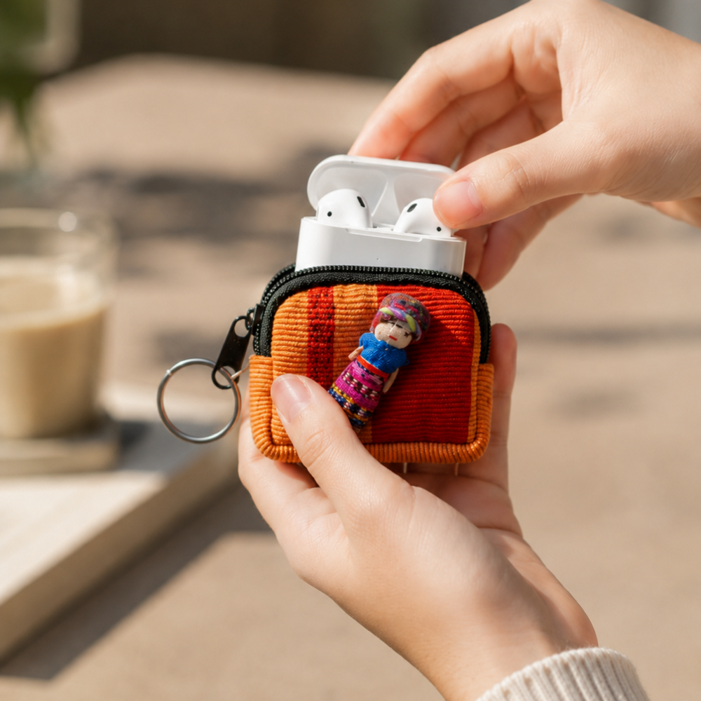 A person holds a small zippered fabric AirPods case with a black zipper, orange and red striped woven body, a metal key ring, and a small doll charm. White AirPods are visible inside the open case, and the background shows a softly blurred outdoor setting with a drink.