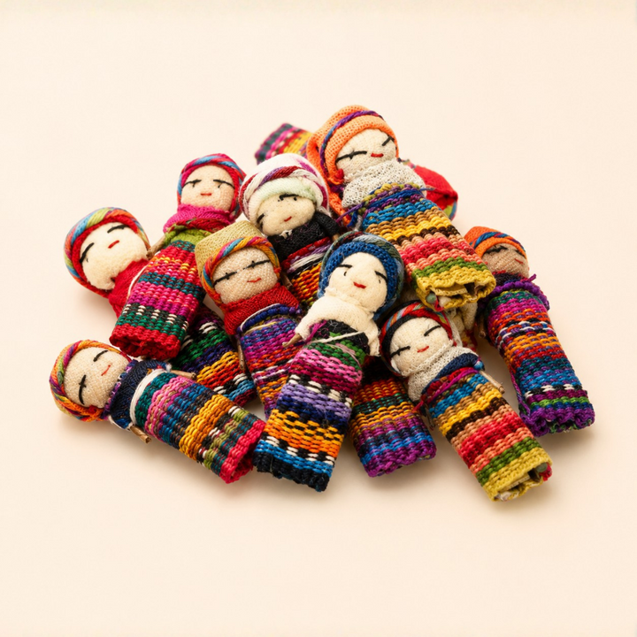 A pile of colorful handmade Guatemalan woven worry doll toys with embroidered faces, arranged on a plain light beige background.