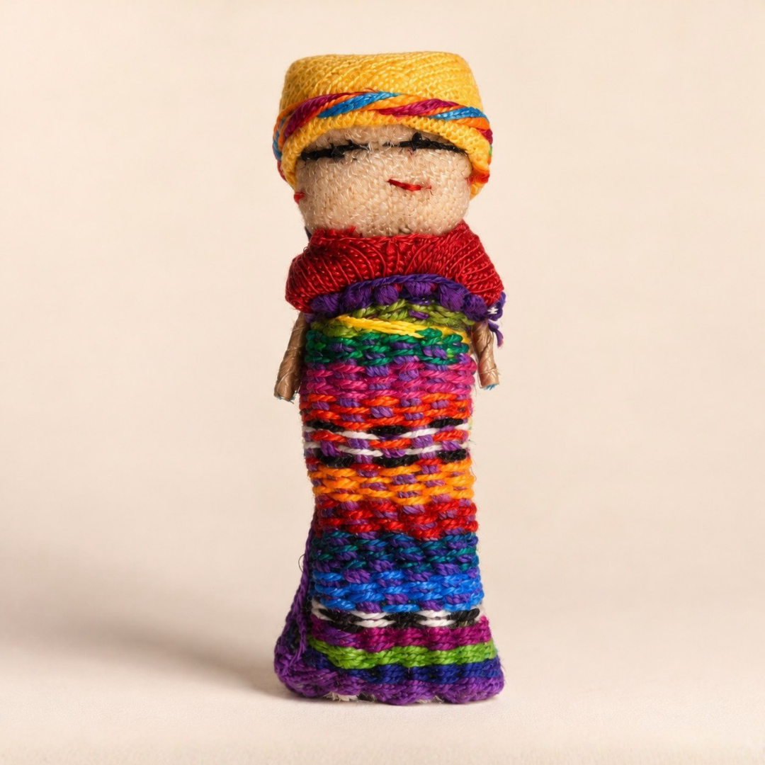 A handmade knitted guatemalan worry doll with a yellow striped hat, red scarf, and a long multicolored striped body, standing against a plain beige background.
